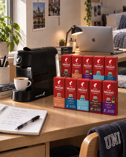 A wooden desk in a cozy home office setting displays a collection of Julius Meinl Coffee Nespresso boxes in various flavors alongside a black Julius Meinl coffee machine, called Cino Pinta, and a cup of espresso. The background shows a blurred bedroom and desk lamp, creating a warm, aspirational lifestyle scene.