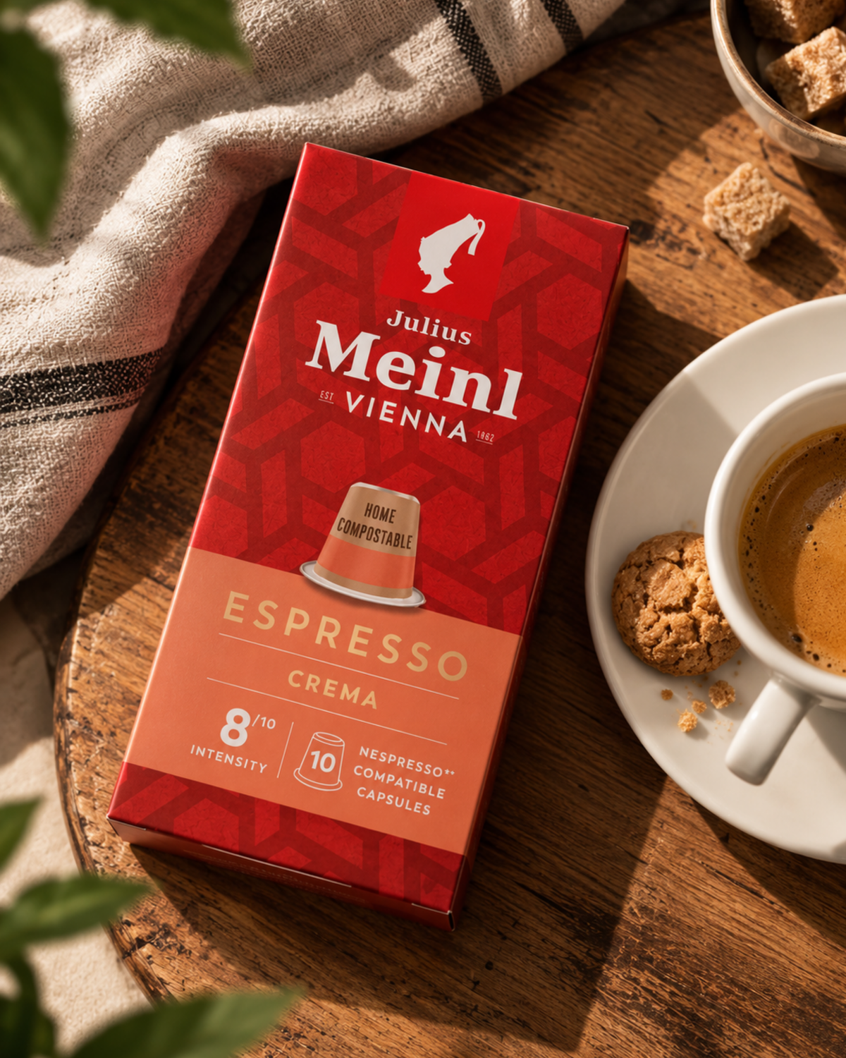 Julius Meinl Vienna Espresso Crema capsule box in red packaging, styled on a wooden table next to a cup of espresso and cookies.
