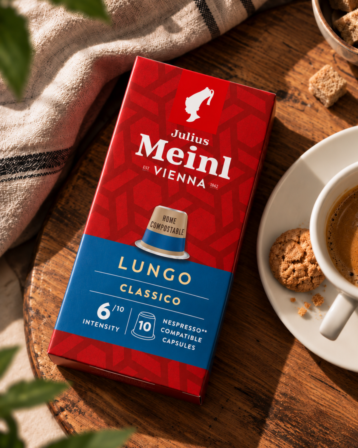 Julius Meinl Vienna Lungo Classico coffee capsule box in red and blue packaging, styled on a wooden table next to a cup of coffee, a cookie, and a striped cloth.