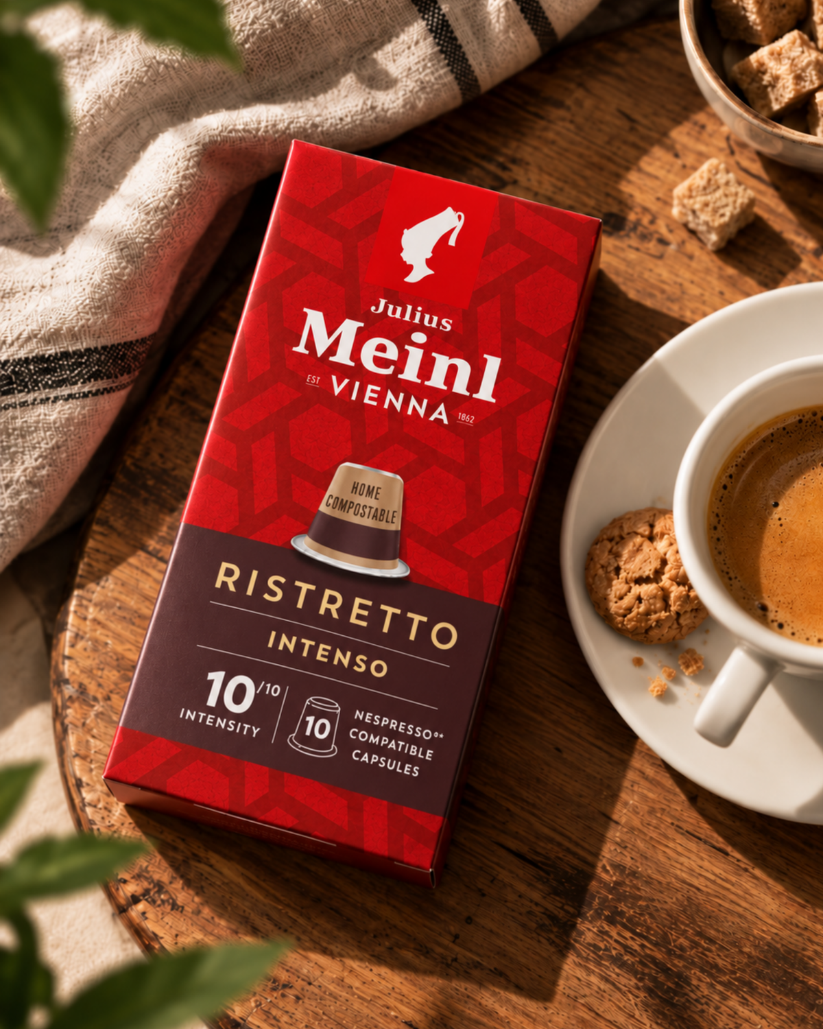A red and dark brown box of Julius Meinl Vienna Ristretto Intenso Nespresso compatible capsules, styled on a wooden table next to a cup of espresso and cookies.